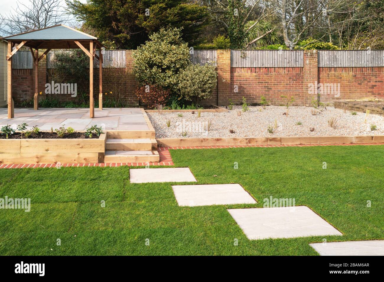 New stepping stones leading to steps and a raised patio with a wooden gazebo, next to a gravel stone planting area. The new turf has a brick mowing st Stock Photo