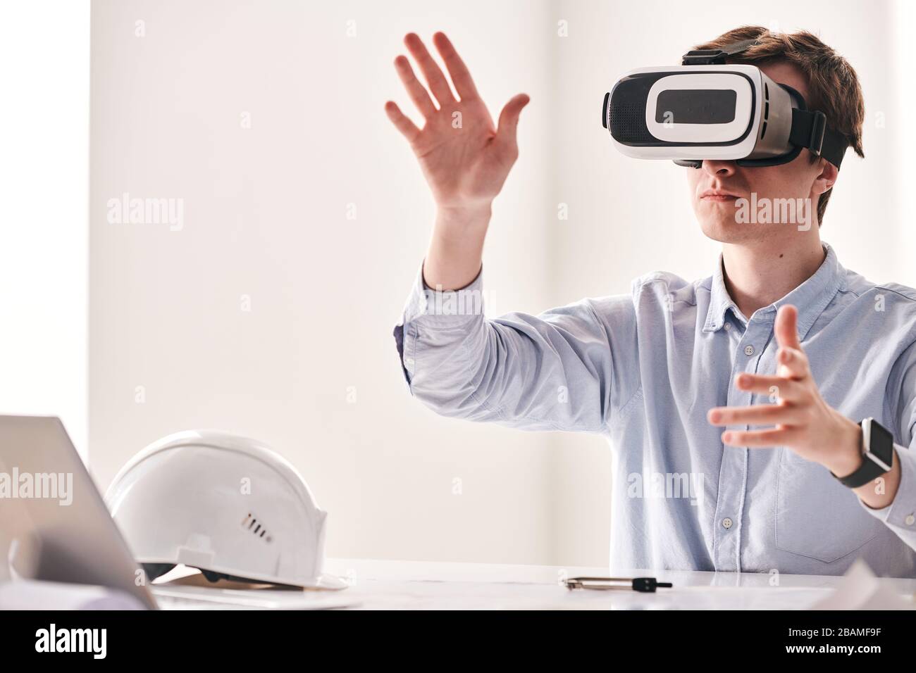 Concentrated young engineer sitting at table and twisting hands while using virtual reality device to make 3D building project Stock Photo