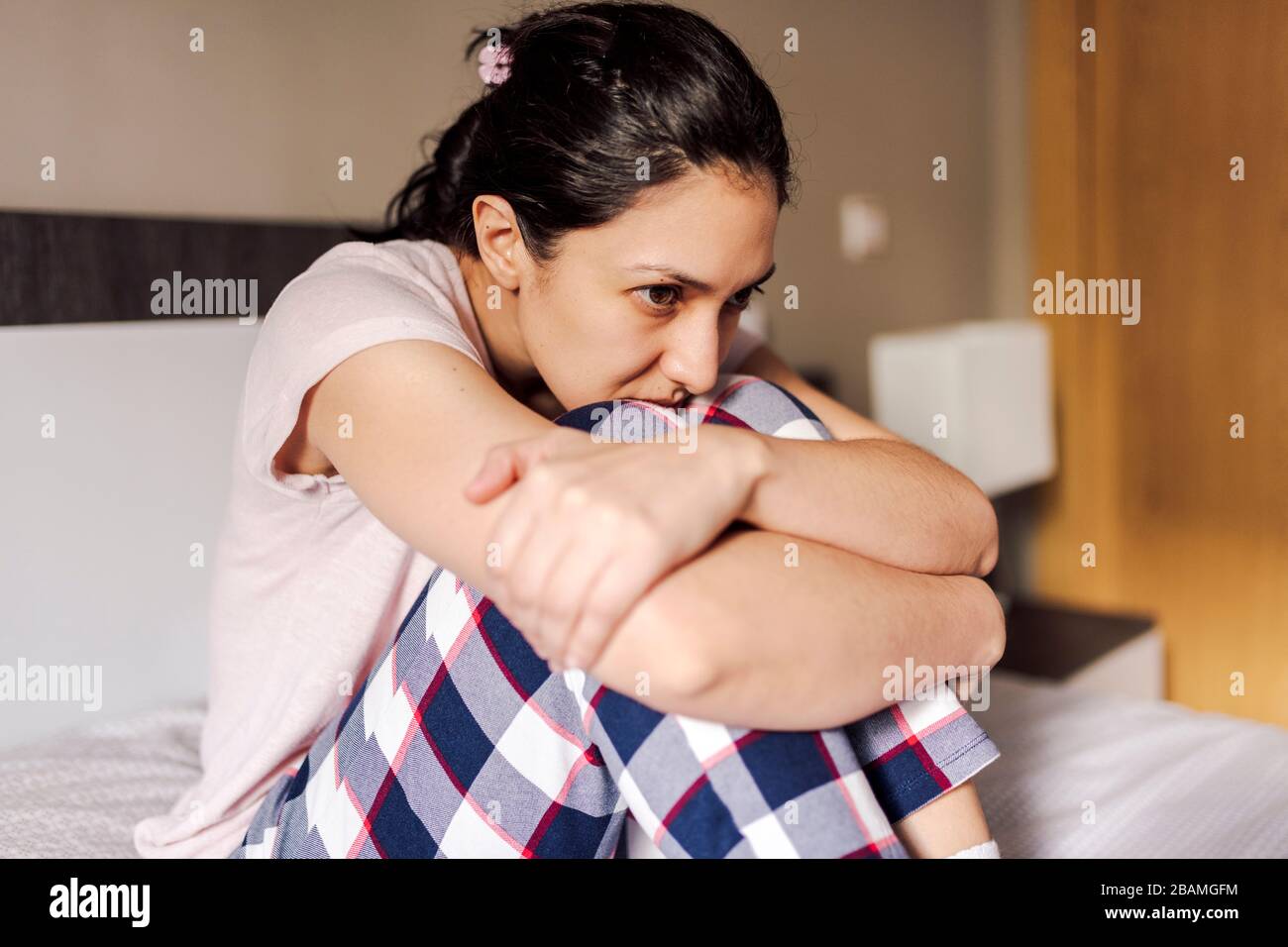 Sad young woman sitting alone in bed in a room. Concept of dramatic loneliness, sadness, depression, sad. Stock Photo