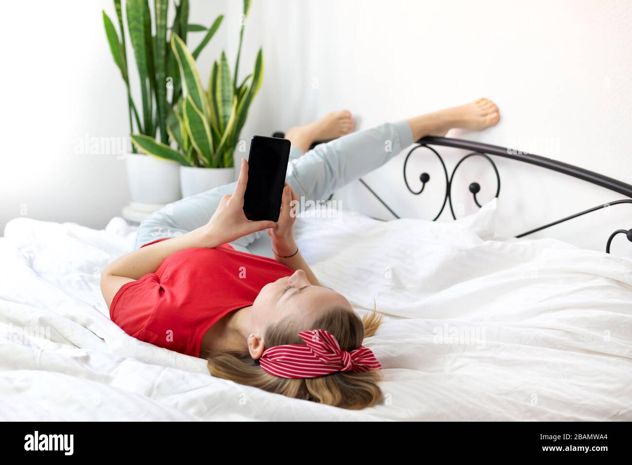 young smiling woman with blond hair in a red T-shirt and comfortable pants lies on a white bed with a smartphone. she works, study from home, writes a Stock Photo