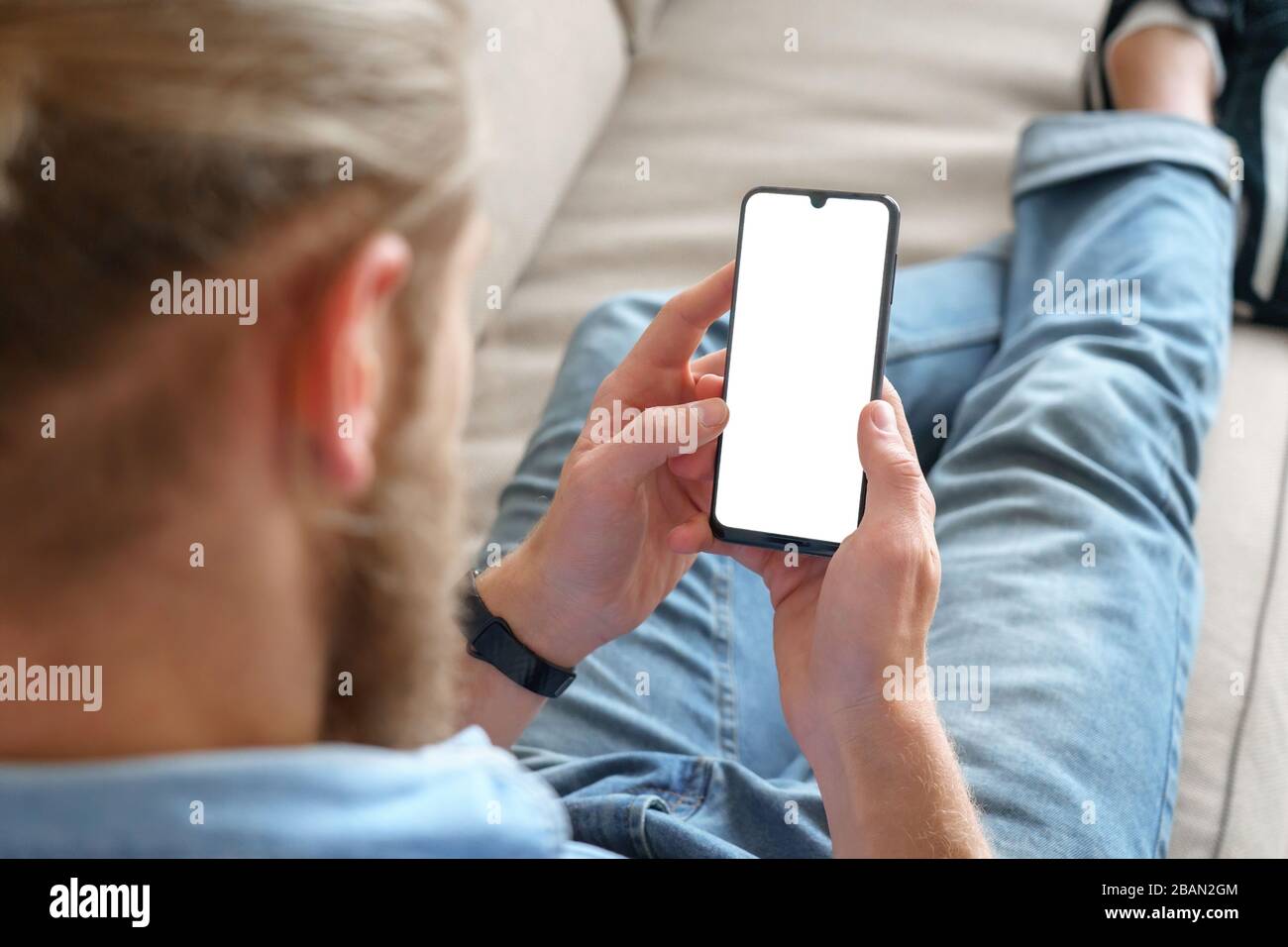 Young male tech user relaxing on sofa using smartphone mock up blank white screen. Stock Photo