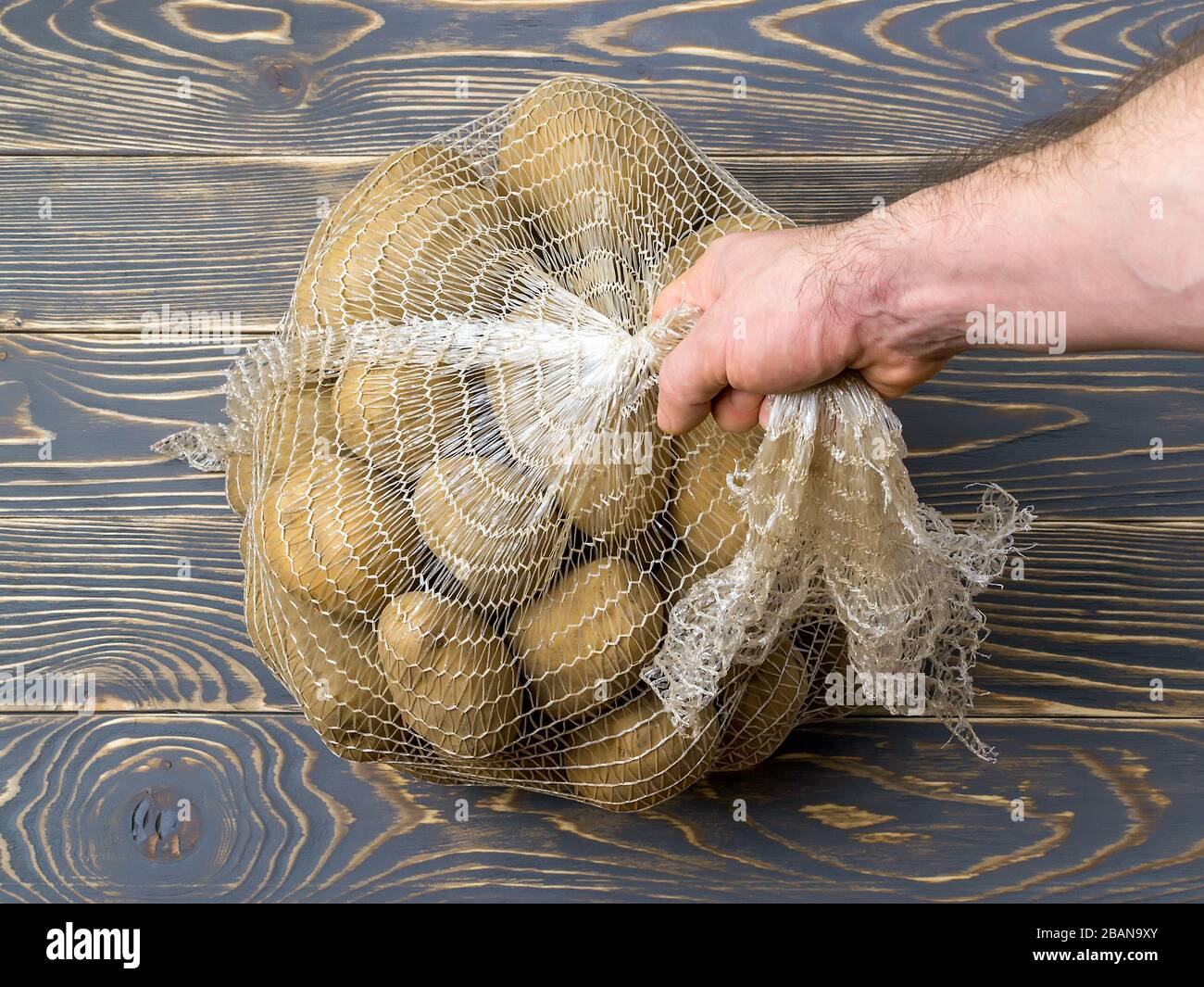 https://c8.alamy.com/comp/2BAN9XY/man-hand-lifts-the-pp-mesh-bag-with-10-kg-of-farm-potatoes-polypropylene-net-sack-with-22-lb-of-organic-potatoes-on-a-dark-wooden-floor-2BAN9XY.jpg