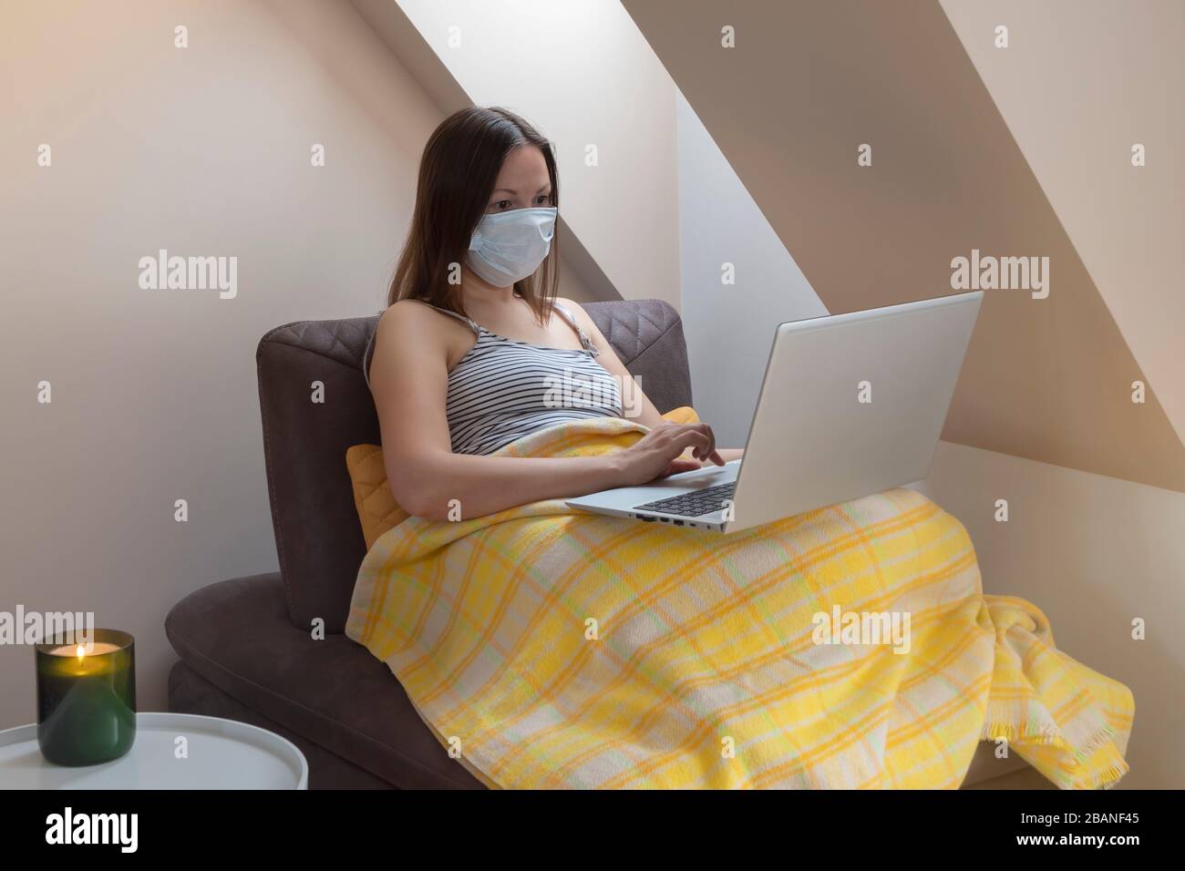 Young woman wearing medical mask on quarantine holding laptop sitting on sofa by the window. Stay at home concept Stock Photo