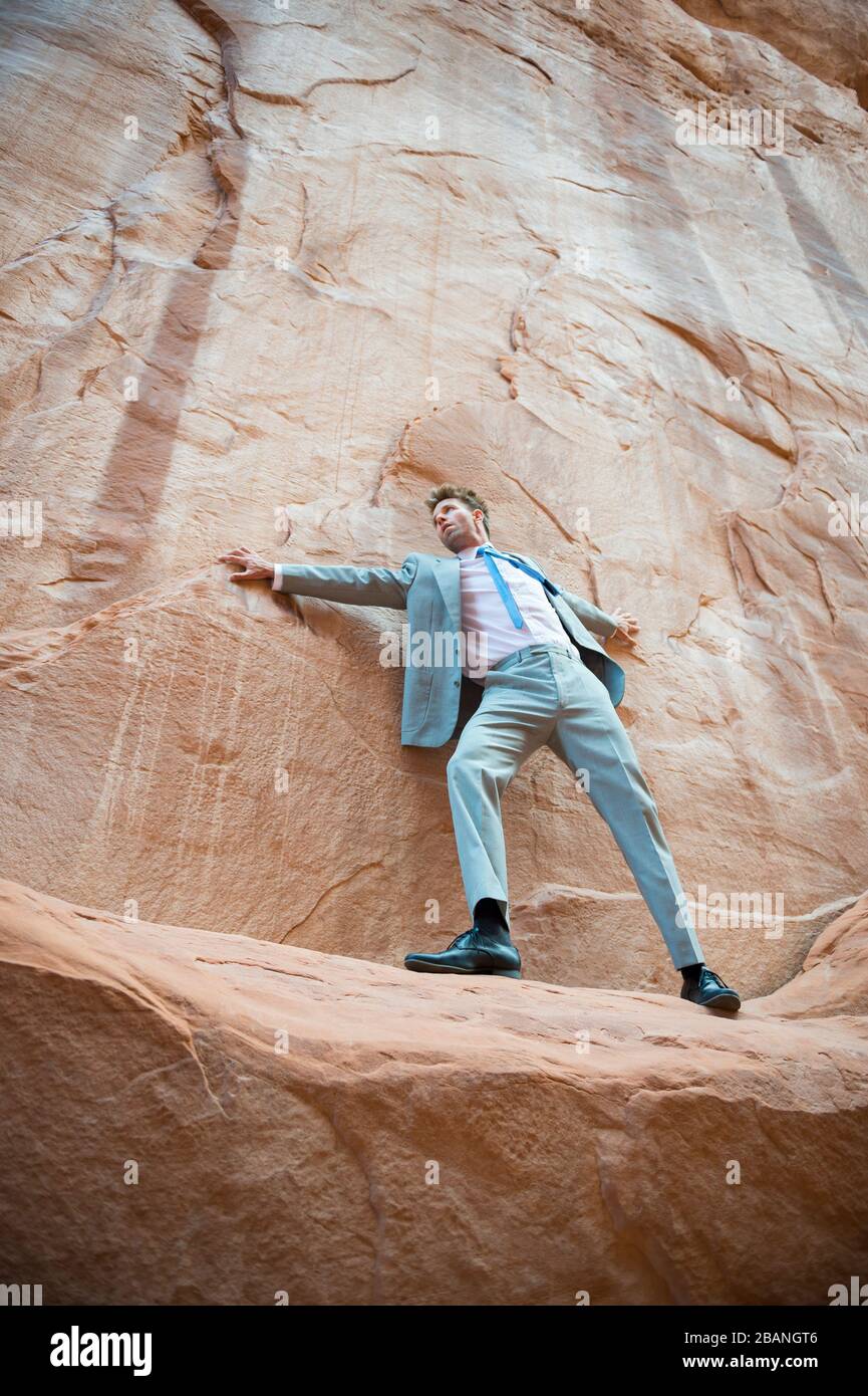 Nervous businessman clinging to a cliff face while balancing on a narrow ledge in a red rock canyon Stock Photo