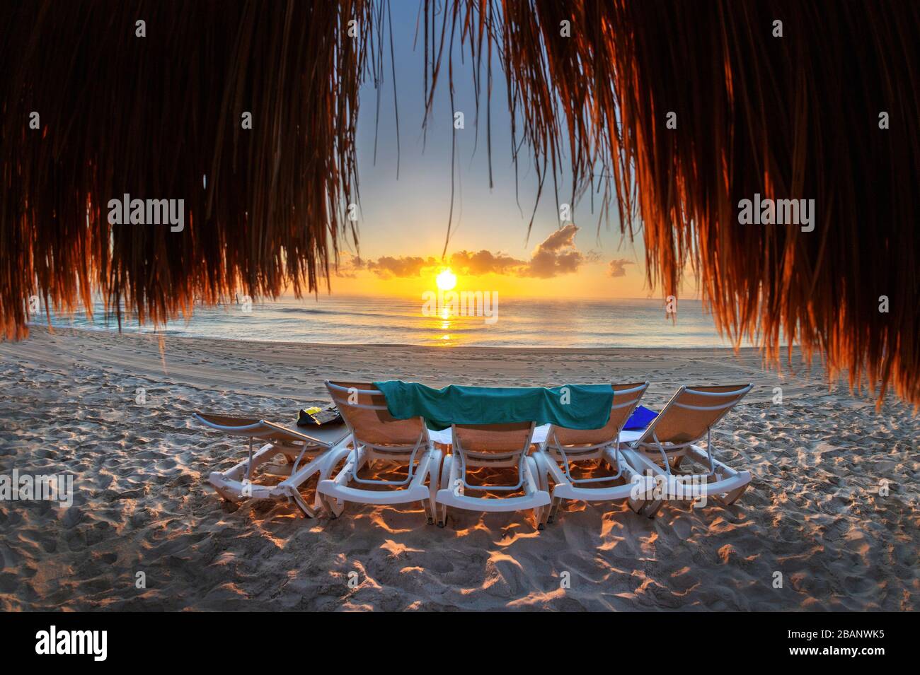 Beautiful golden sunrise over an empty beach and row of lounge chairs on the Caribbean coast of Mexico at Riviera Maya in Cancun as viewed from behind Stock Photo