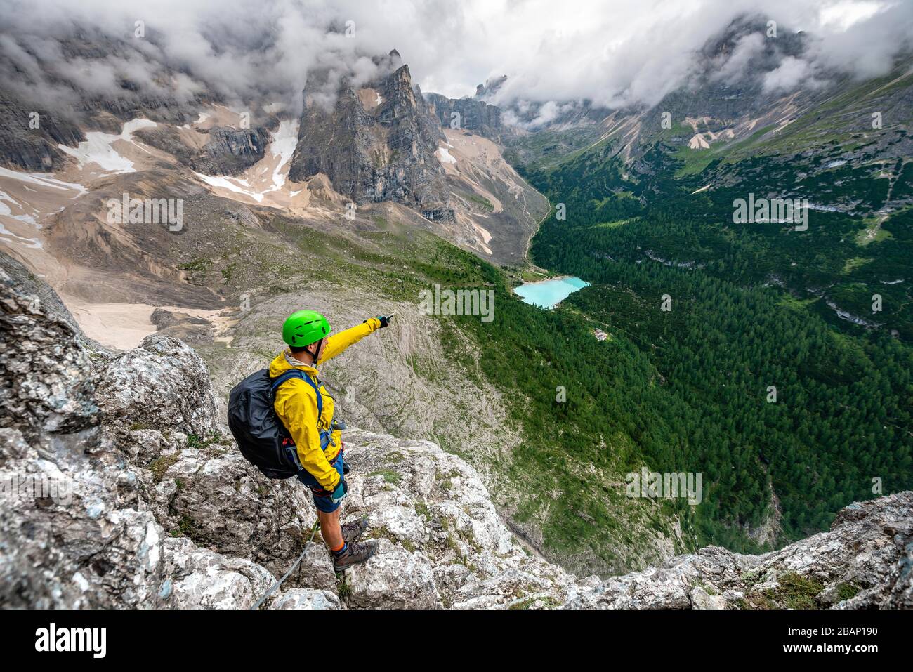 Young man points into the distance, mountaineer on a via ferrata Vandelli, view of Lago di Sorapis, Sorapiss circuit, mountains with low clouds Stock Photo