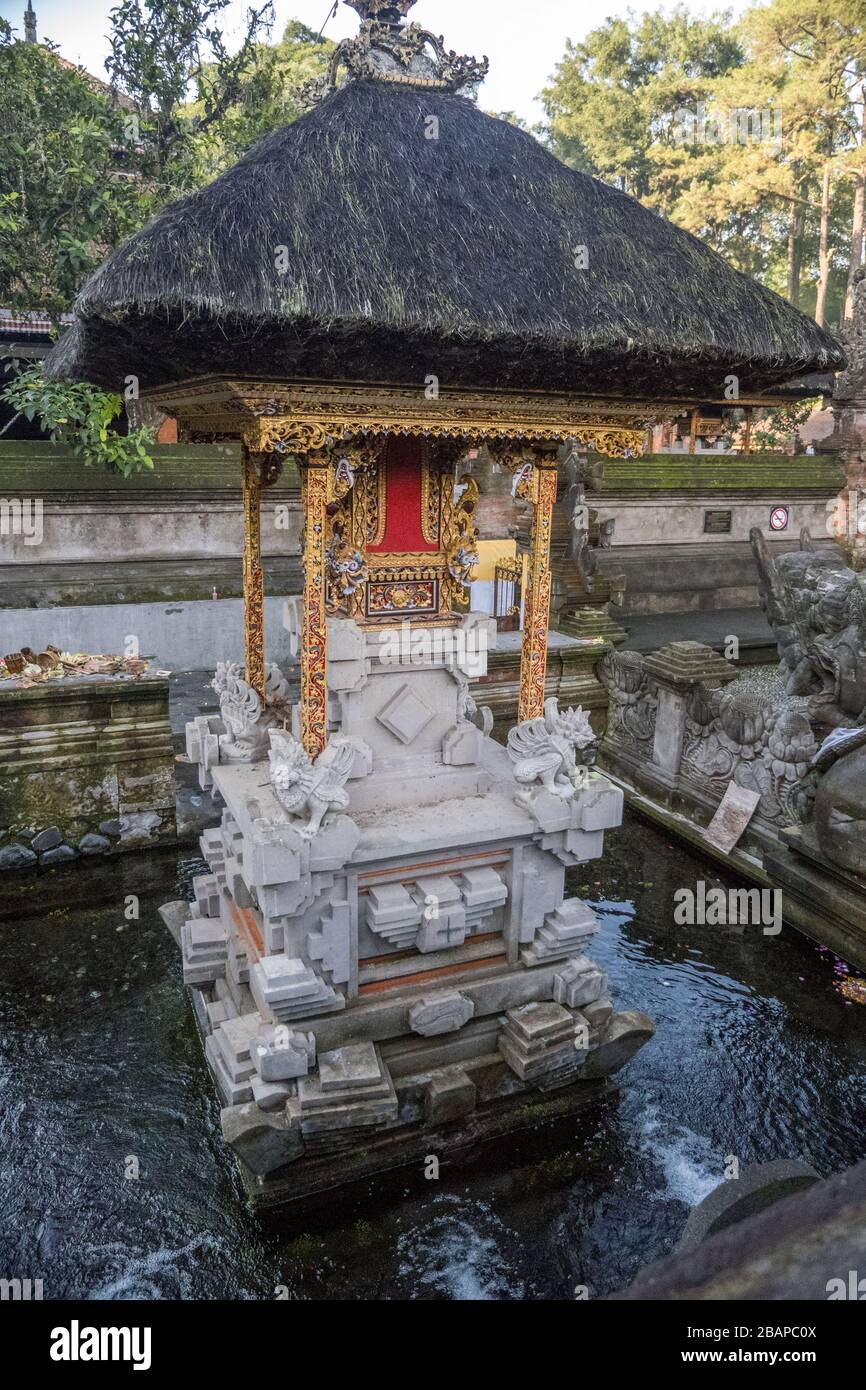 Small stone temple sitting in pool of water with golden frame, bright red clothe and thick black thatch roof in Tirta Empul Holy Water Temple Bali. Stock Photo