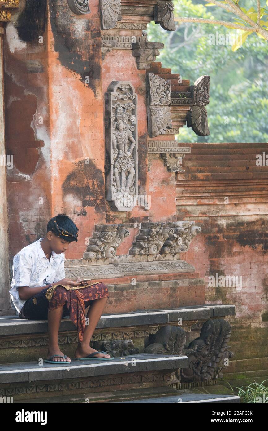 Young Balinese boy sitting on ornately carved, red Hindu temple steps in early morning looking at his cell phone. Stock Photo