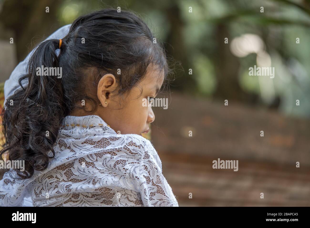 Portrait of little Balinese girl wearing white lace top, black hair pulled back sitting on father's shoulder looking intently into space. Stock Photo
