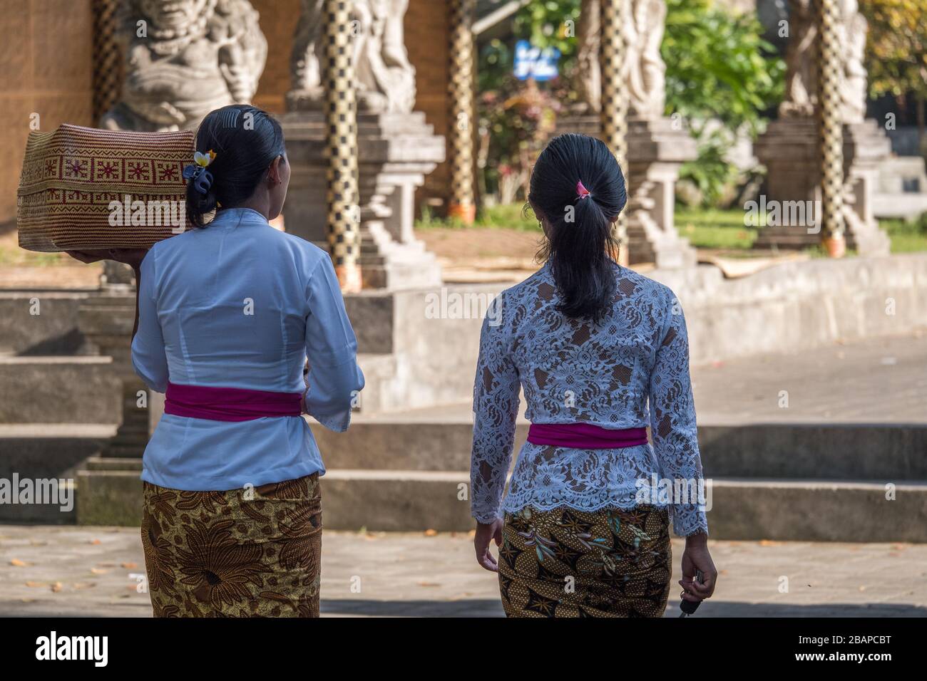 Two young female Holy Water Temple employees dressed in traditional gold sarong, red sash and white lace top carrying woven basket in early morning. Stock Photo