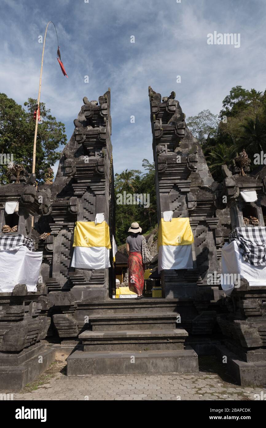 Woman dressed in straw hat, red and black sarong and black top walking up carved stone temple entrance steps in early morning with blue sky. Stock Photo