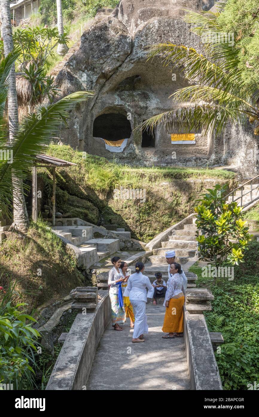 Three Balinese Hindu women dressed in traditional sarong standing and talking on temple bridge with temple buildings and palm trees in background. Stock Photo