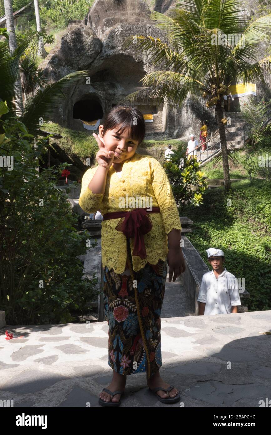 Young Balinese girl in bright yellow top and floral skirt looking at camera with big smile and peace symbol on raised left hand with temple in back. Stock Photo