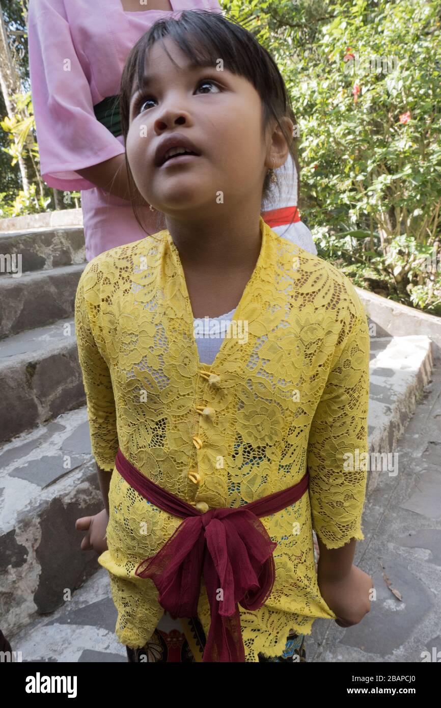 Young Balinese girl in bright yellow top with red sash belt inquisitively looking up at Mother on steps of Hindu Temple. Stock Photo