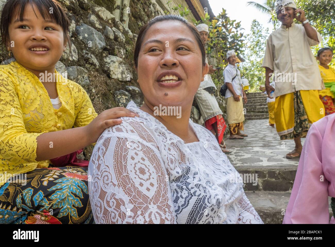 Balinese mother wearing white lace top, smiling, young daughter resting hand on her shoulder, sitting on temple wall with Father in background. Stock Photo