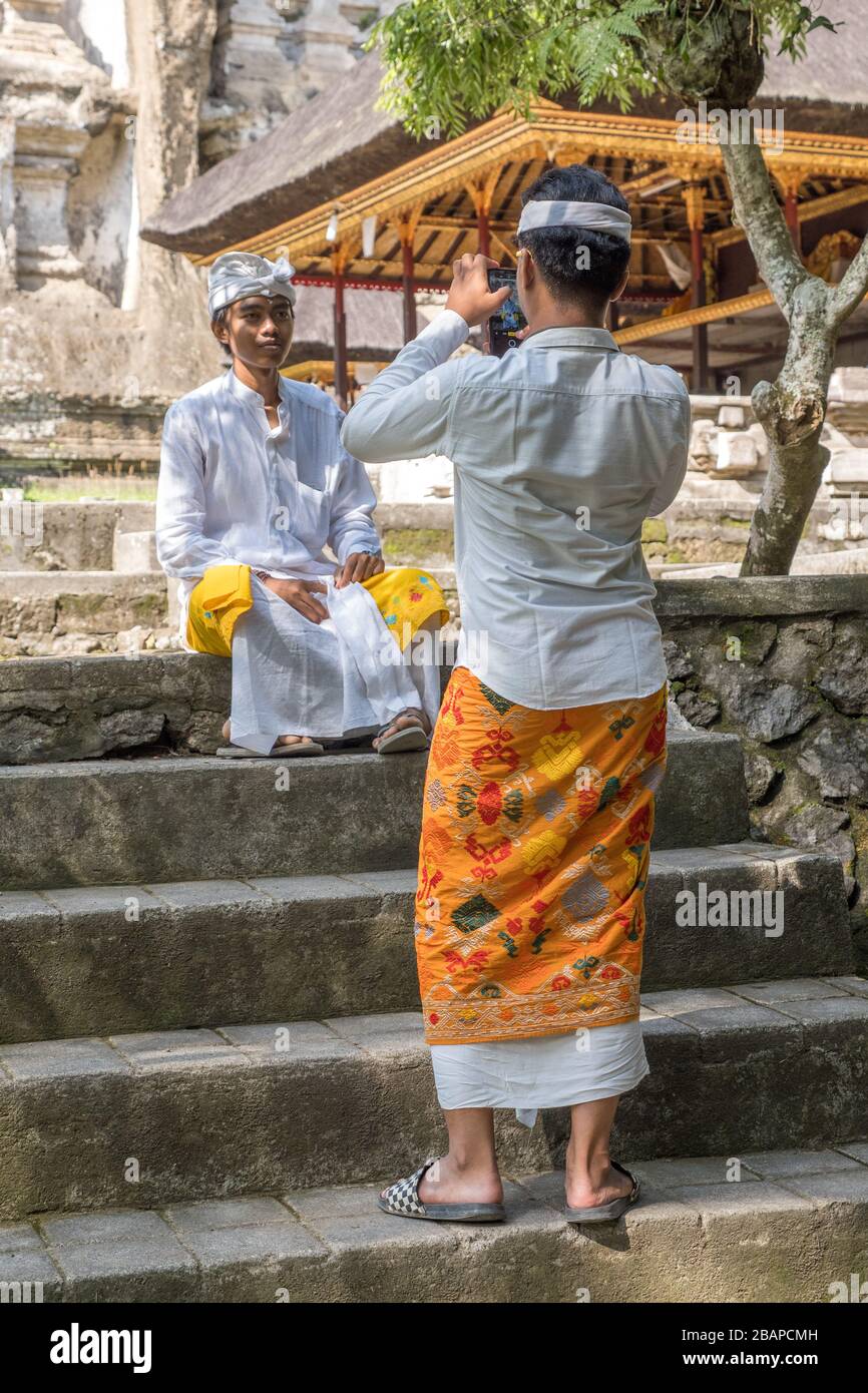 Balinese teenager dressed in traditional white clothing sitting on temple steps posing for photo taken by friend dressed in colorful sarong and udeng. Stock Photo