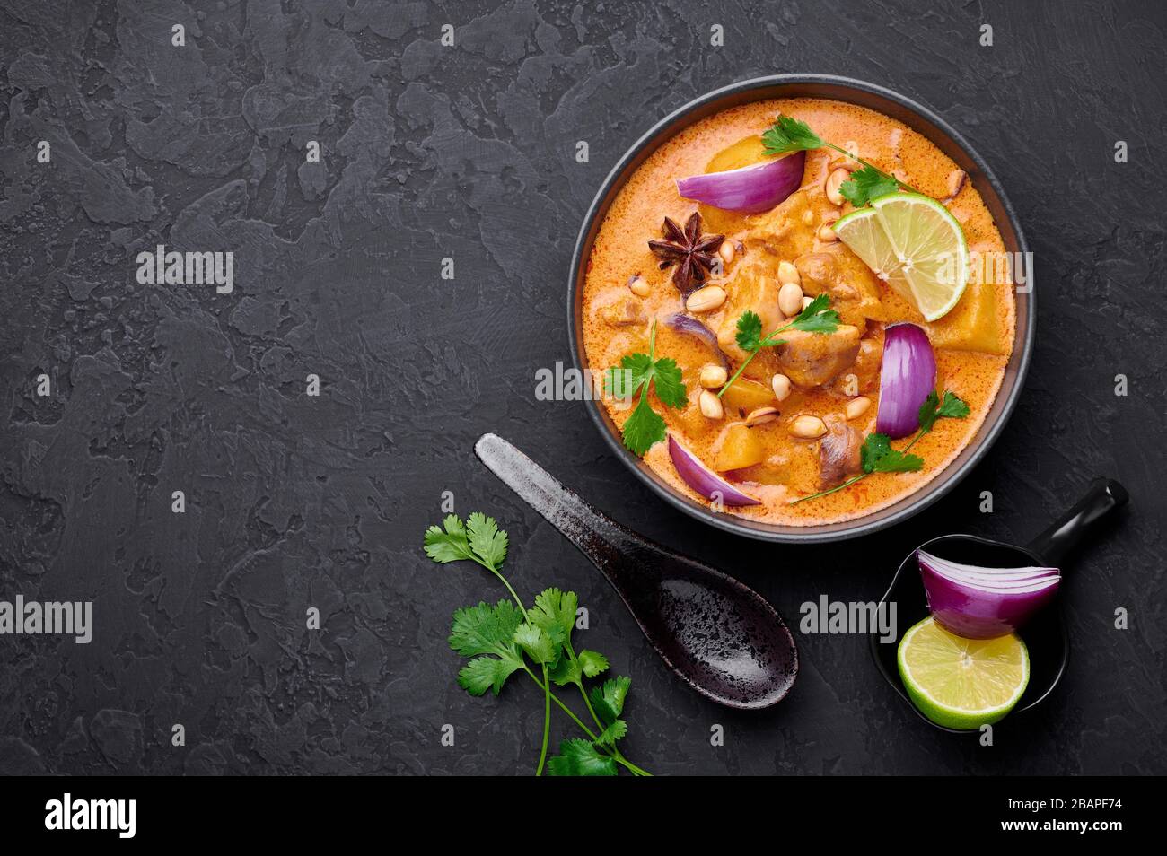 A Chicken Massaman Curry in black bowl at dark slate background. Massaman Curry is Thai Cuisine dish with chicken meat, potato, onion and many spices. Stock Photo