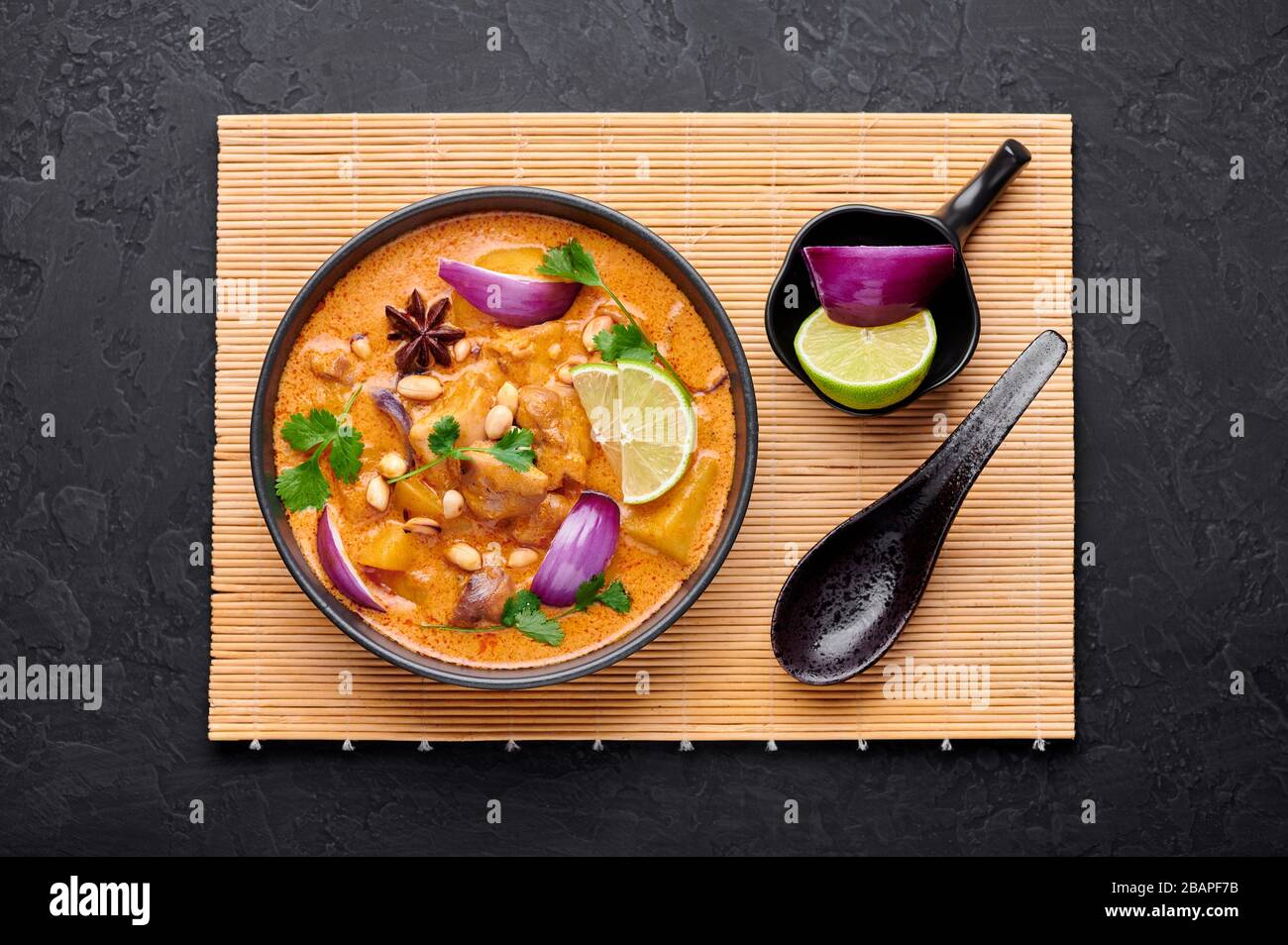 A Chicken Massaman Curry in black bowl at dark slate background. Massaman Curry is Thai Cuisine dish with chicken meat, potato, onion and many spices. Stock Photo