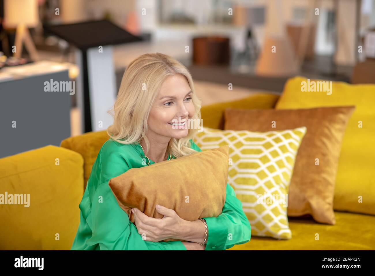 Good mood. Blonde woman in a green blouse huggoing the pillow and smiling Stock Photo