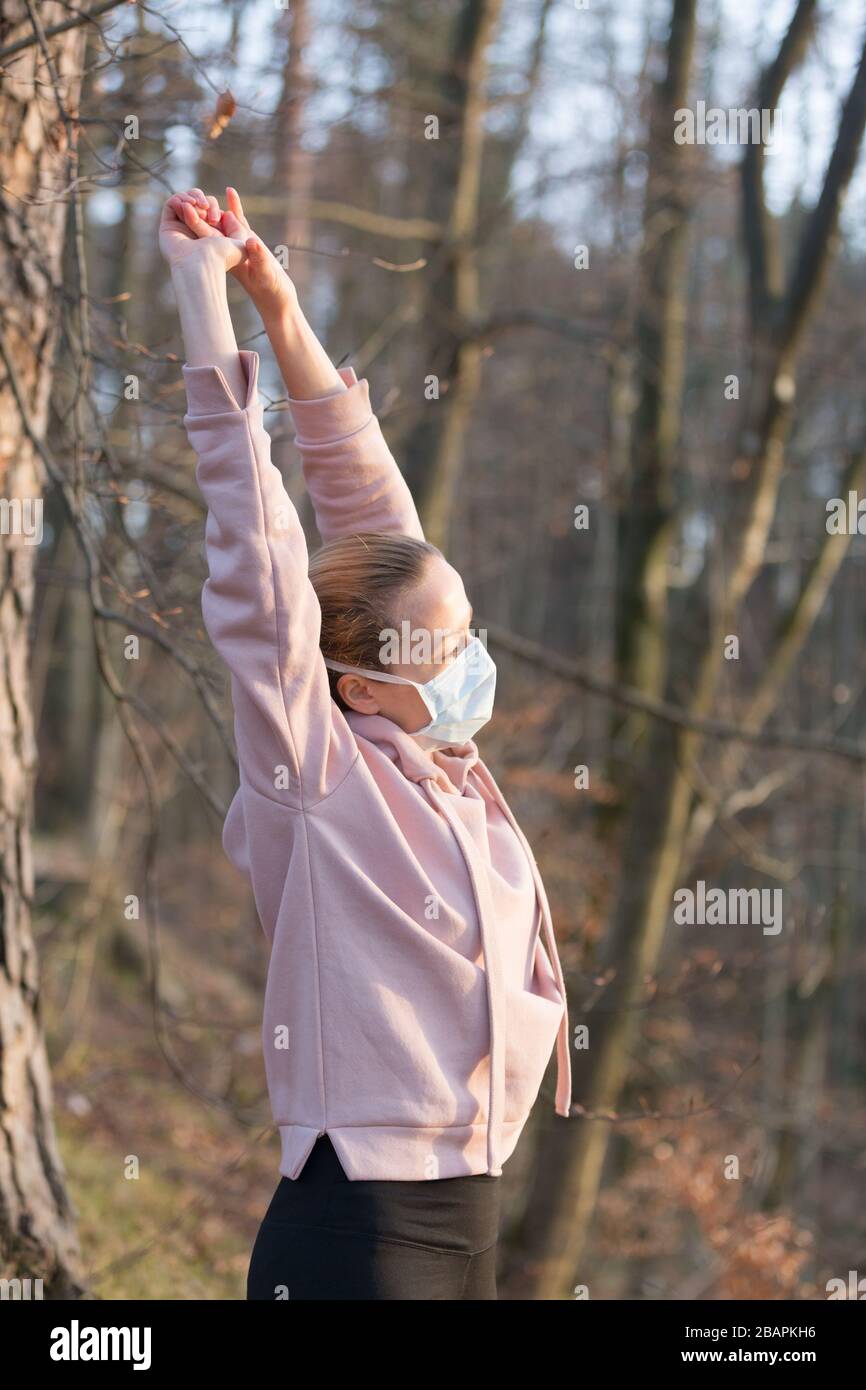Portrait of caucasian sporty woman wearing medical protection face mask while relaxing by stretching in forest. Corona virus, or Covid-19, is Stock Photo