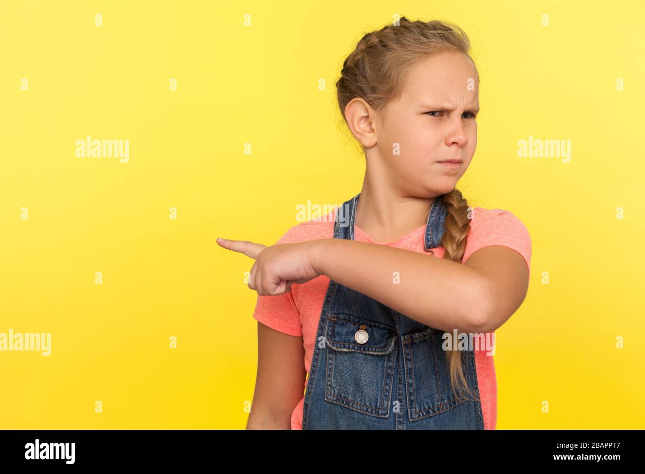Get out! Portrait of angry upset little girl with braid in denim overalls pointing way out showing exit, asking to leave her alone, feeling resentful. Stock Photo