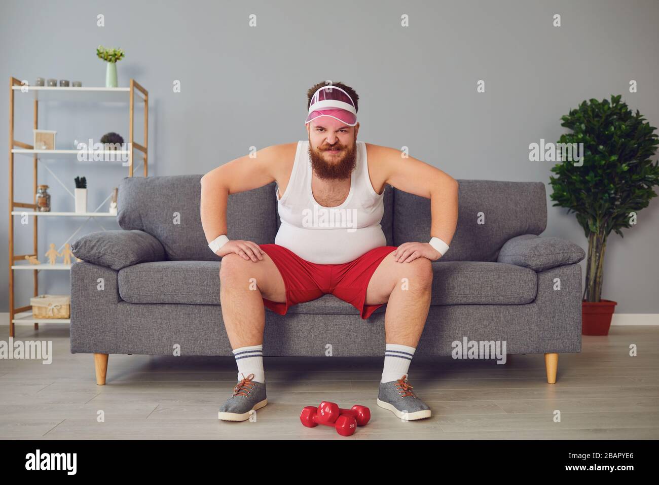 Funny fat angry man in sportswear is sitting on the sofa in the room Stock Photo