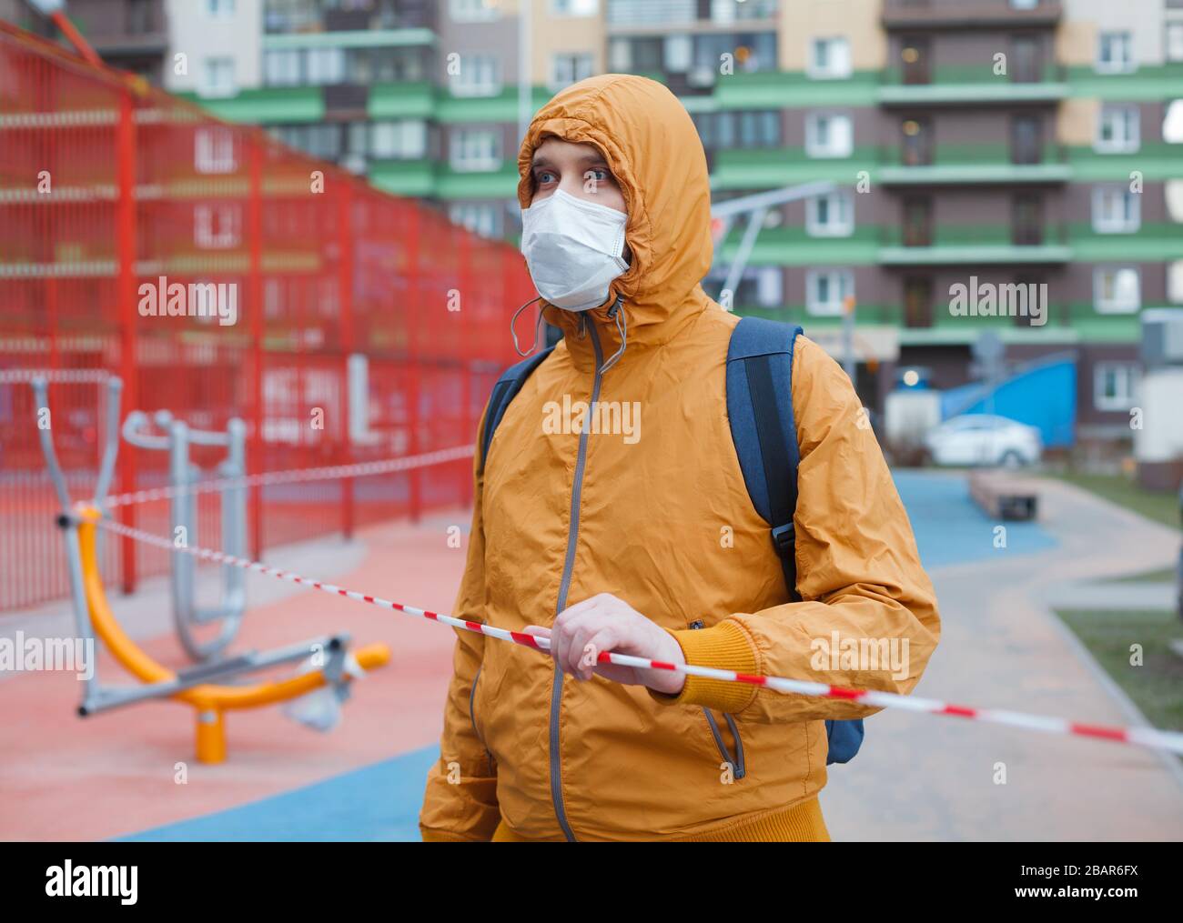 Man in hood and medicine mask stand in his yard touching tape fencing outdoor gym. Coronavirus quarantine. Stock Photo