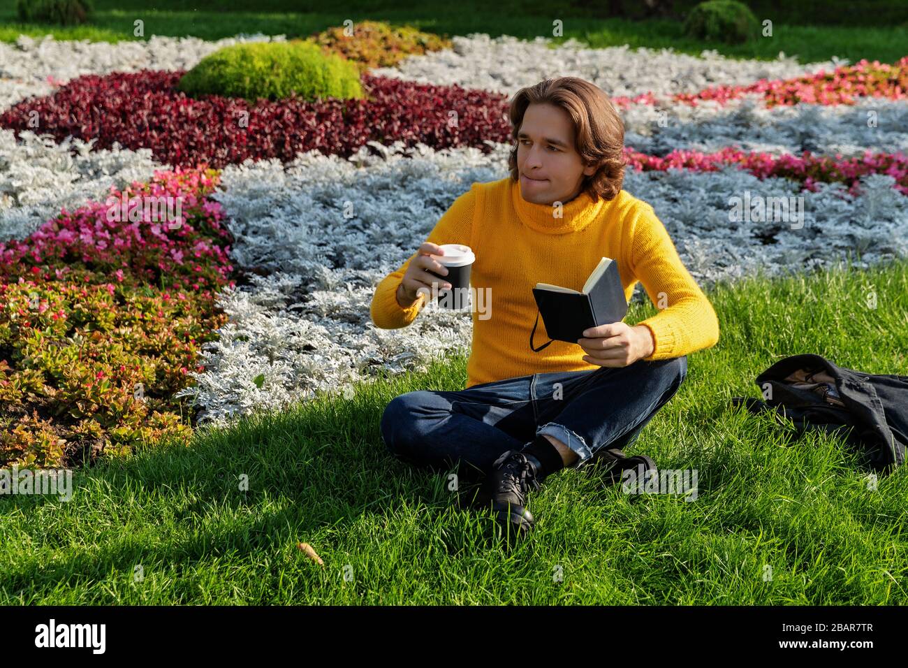 The young man drinks coffee and reads the book in the park, he smiles, has a rest, is dressed in a yellow sweater, flowers and grass on background Stock Photo