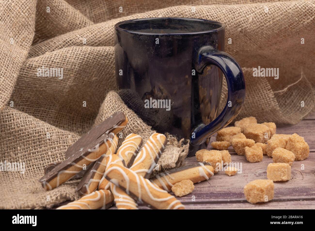dark blue ceramic tea mug, cookie sticks with chocolate and white icing and pieces of brown cane sugar on a background of textured fabric. Close up Stock Photo