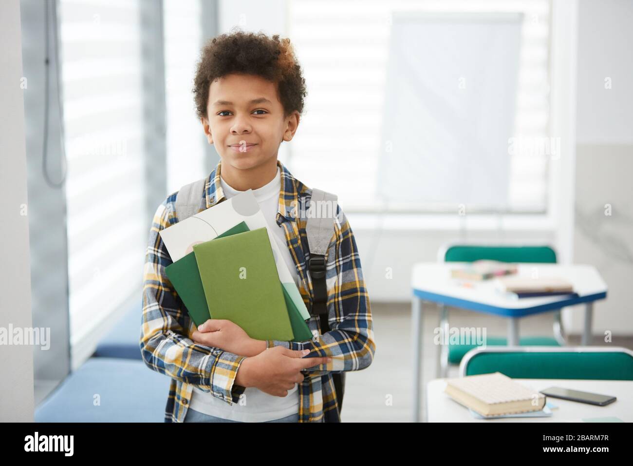 Waist up portrait of smiling African-American boy holding books and looking at camera while posing in school classroom, copy space Stock Photo