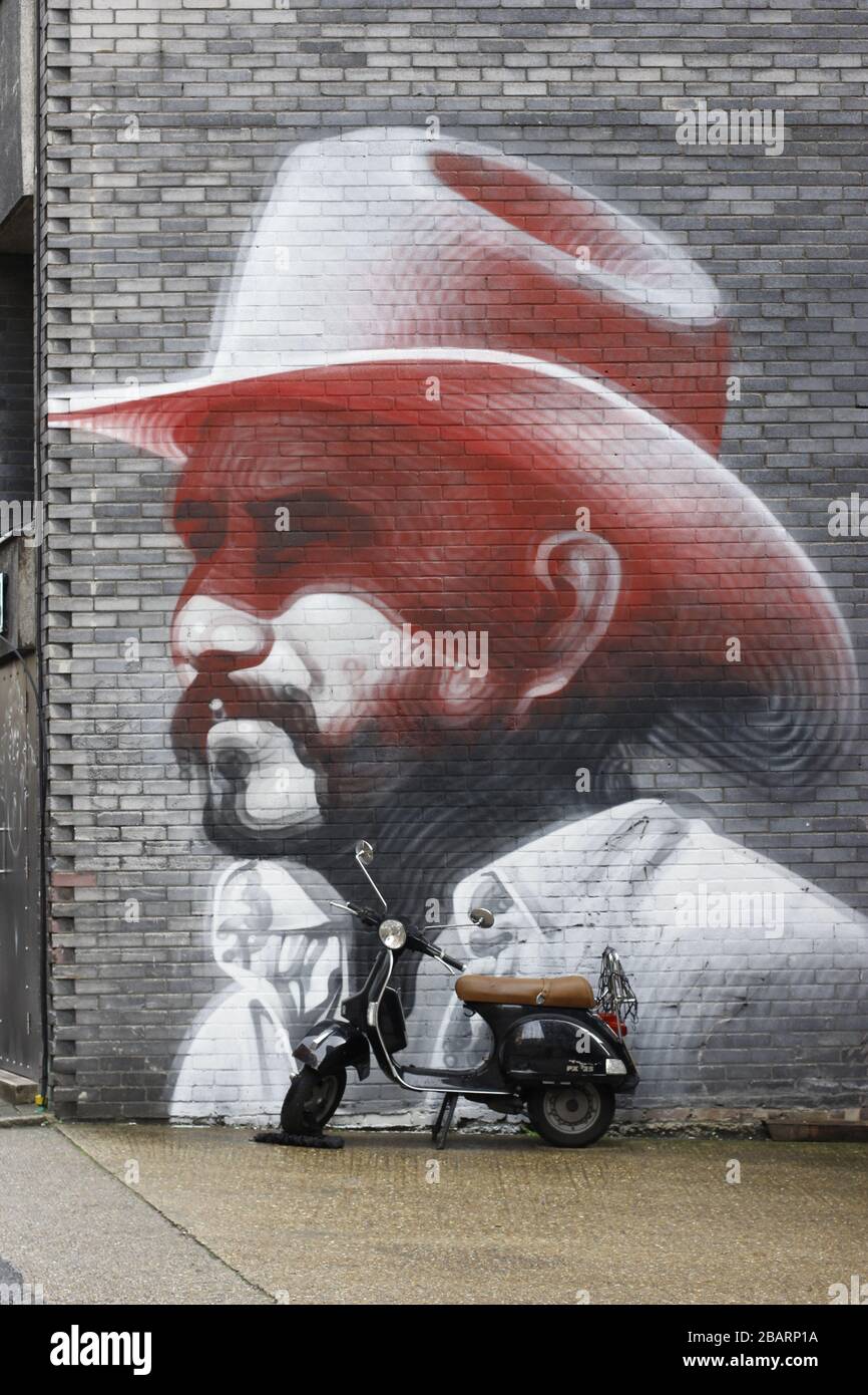 Large scale street artwork on a wall of a building depicting a man wearing a pink cowboy hat. A motor scooter is parked alongside Stock Photo