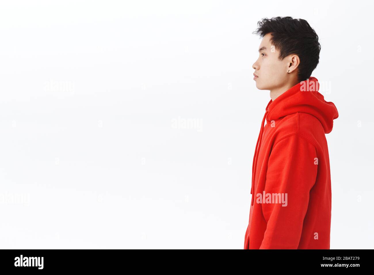 Profile portrait of young asian man in red hoodie looking left with serious, unbothered expression, standing casually over white background, concept Stock Photo