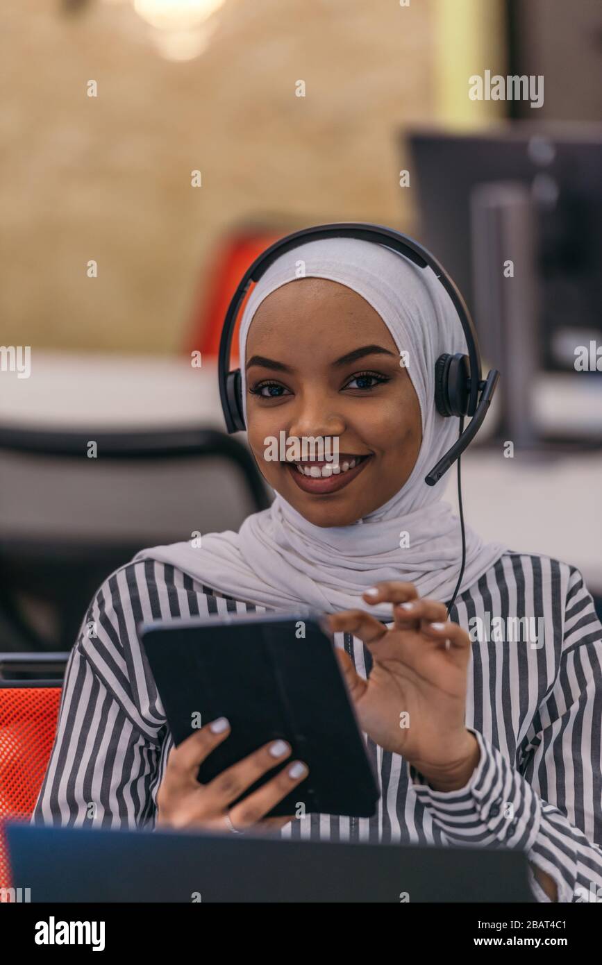 Happy African muslim girl with hijab working on a tablet with a headset ...