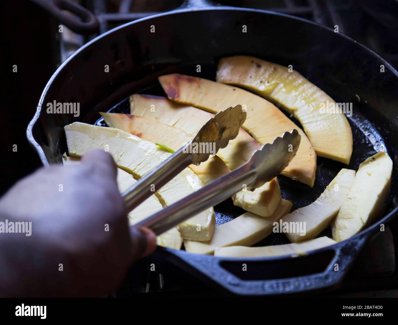 The hand of a black man, holding tongs while frying sliced breadfruit in shallow oil, in a black iron pot on a stove top flame Stock Photo