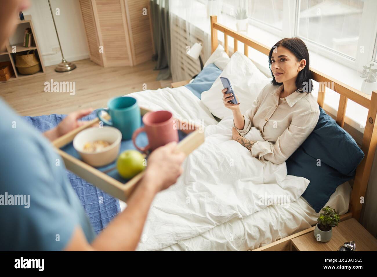 High angle view at unrecognizable caring man bringing breakfast in bed for young woman holding smartphone, warm-toned family scene, copy space Stock Photo