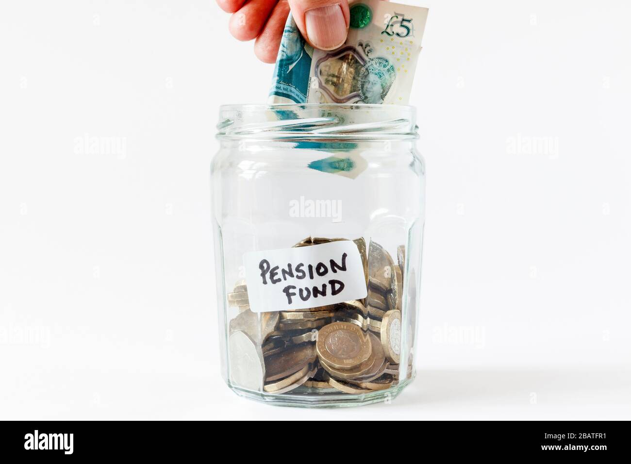 A glass jam jar containing British coins, with a handwritten label 'pension fund', a male hand inserting a £5 note Stock Photo