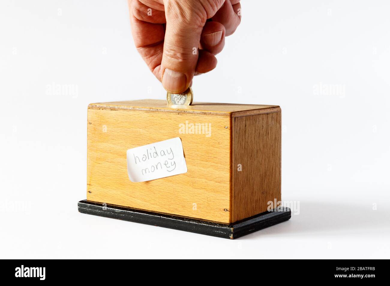 A home-made wooden money box with a handwritten label 'holiday money', a male hand inserting a £1 coin Stock Photo