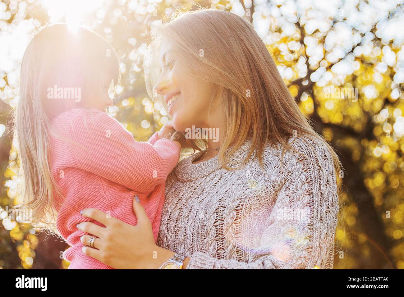 Closeup of happy young family spending time together outdoors. Young mother holding her little daughter in hands and hugging her while standing in aut Stock Photo