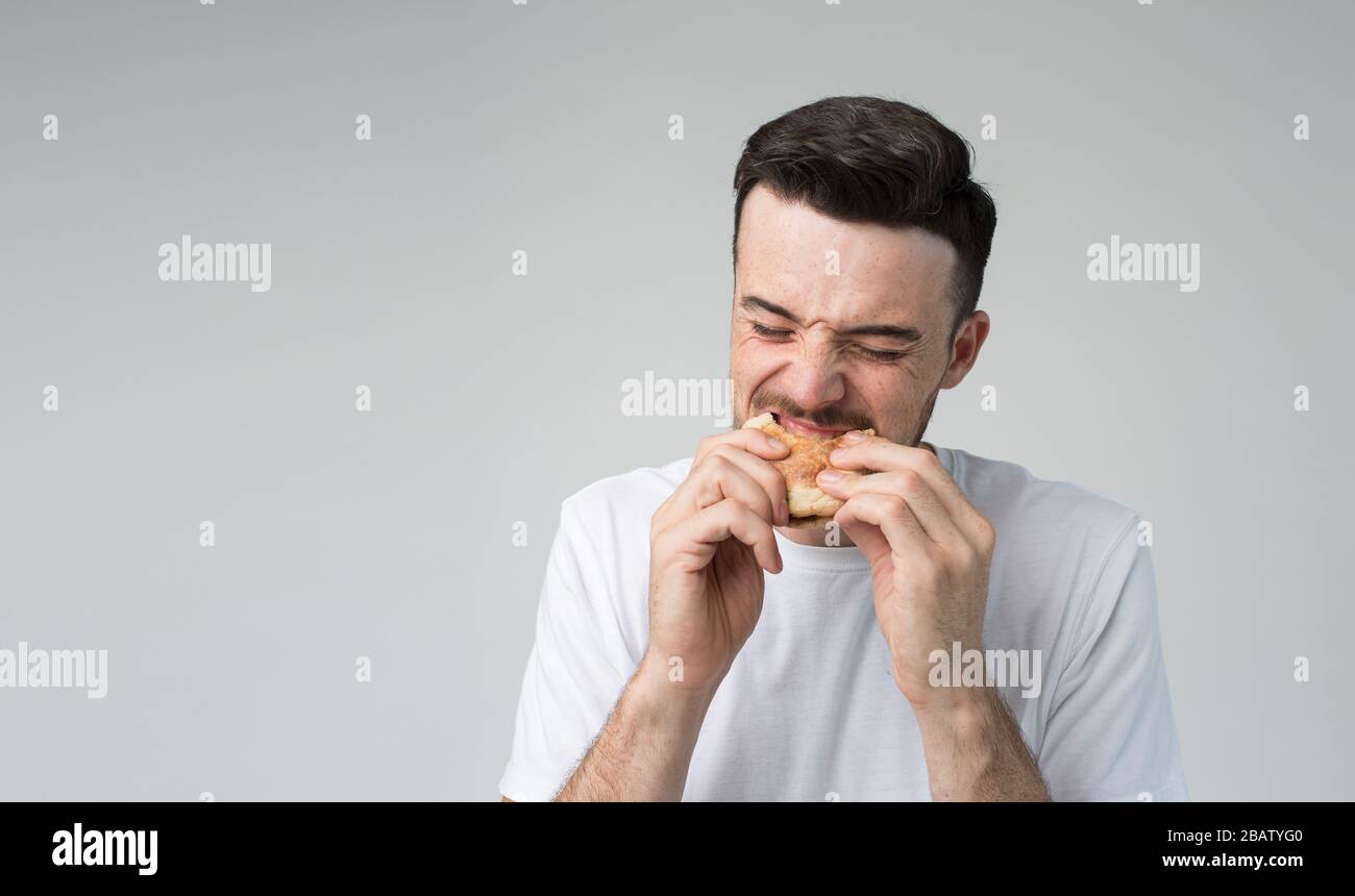 Young man isolated over background. Picture of guy eating burger and biting it. Tasty delisious fatning meal. Eating with pleasure. No guilty for Stock Photo