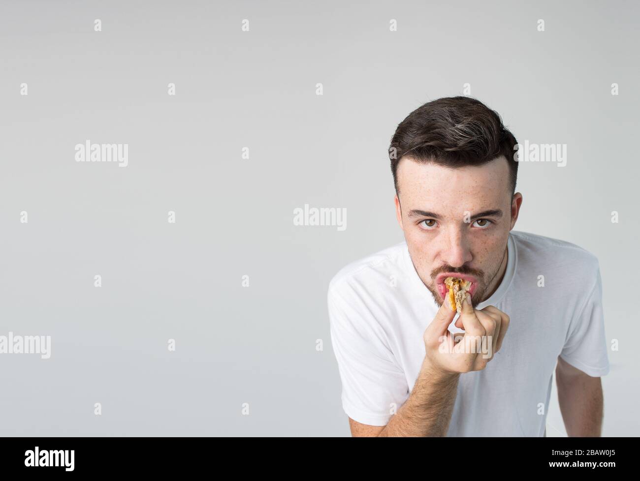 Young man isolated over background. Strange weirdo look straight on camera and biting piece of yummy tasty burger. Guy in white shirt posing alone in Stock Photo