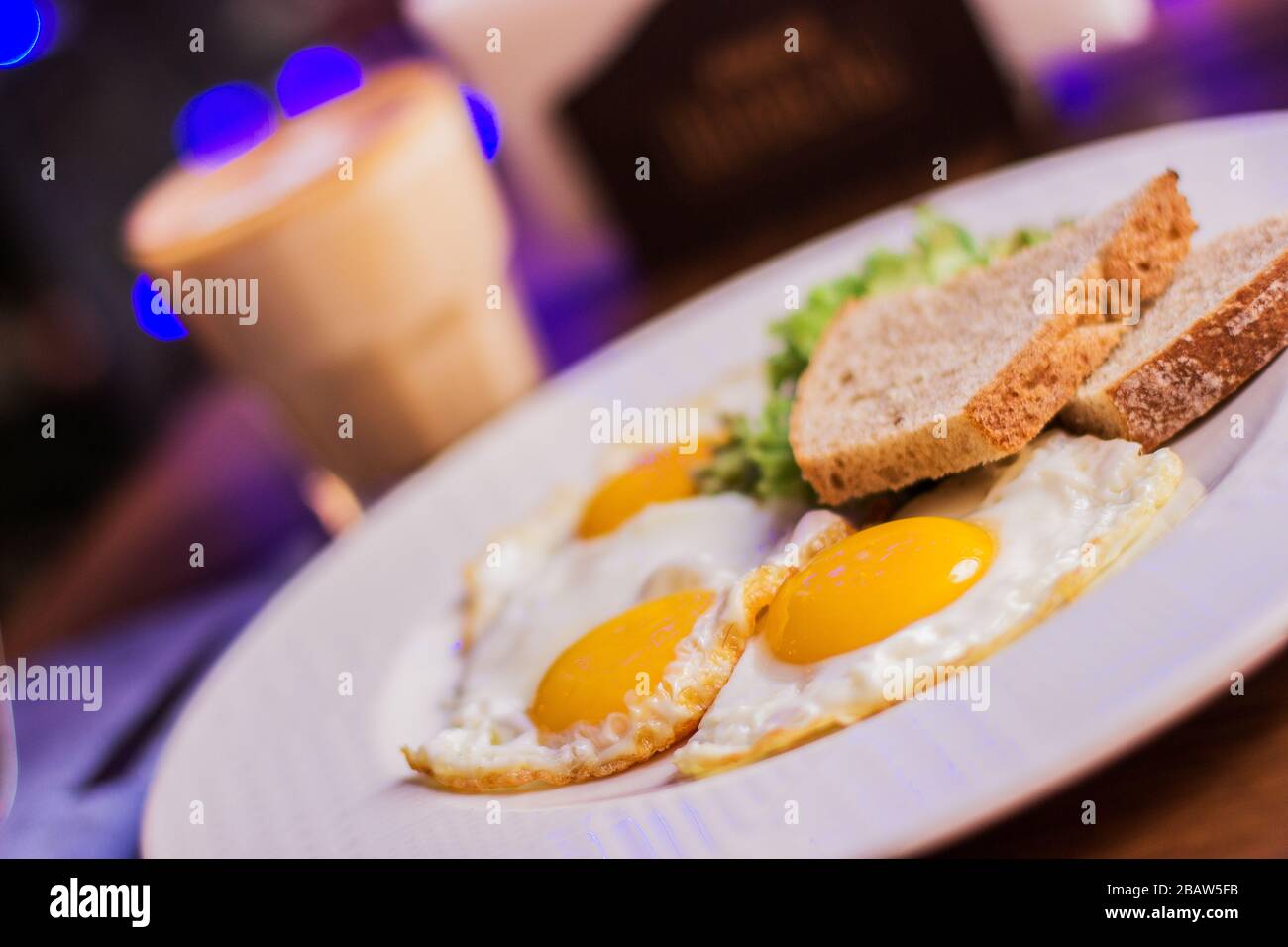 Closeup of fried eggs with toasted bread on white dish and glass with coffee on wooden table at modern restaurant. Traditional american breakfast in d Stock Photo