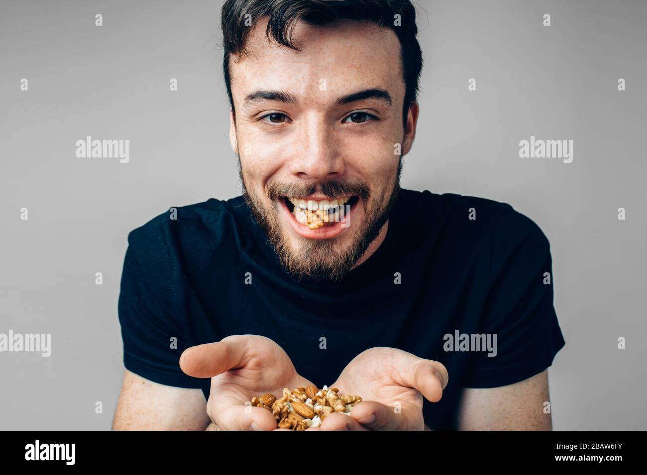 Young man isolated over background. Guy in black clothes hold differents nuts in hands and one wanut between teeth. Healthy eating and lifestyle Stock Photo