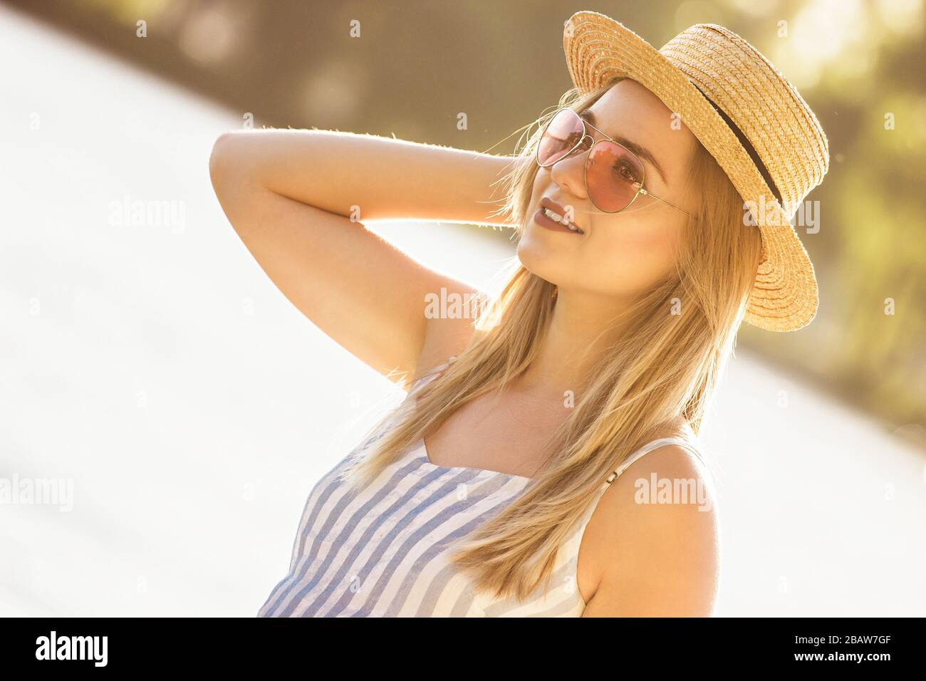 Portrait of tender young woman in straw hat and sunglasses standing at sunny riverside. Beautiful stylish girl in stripped overalls posing at beach on Stock Photo