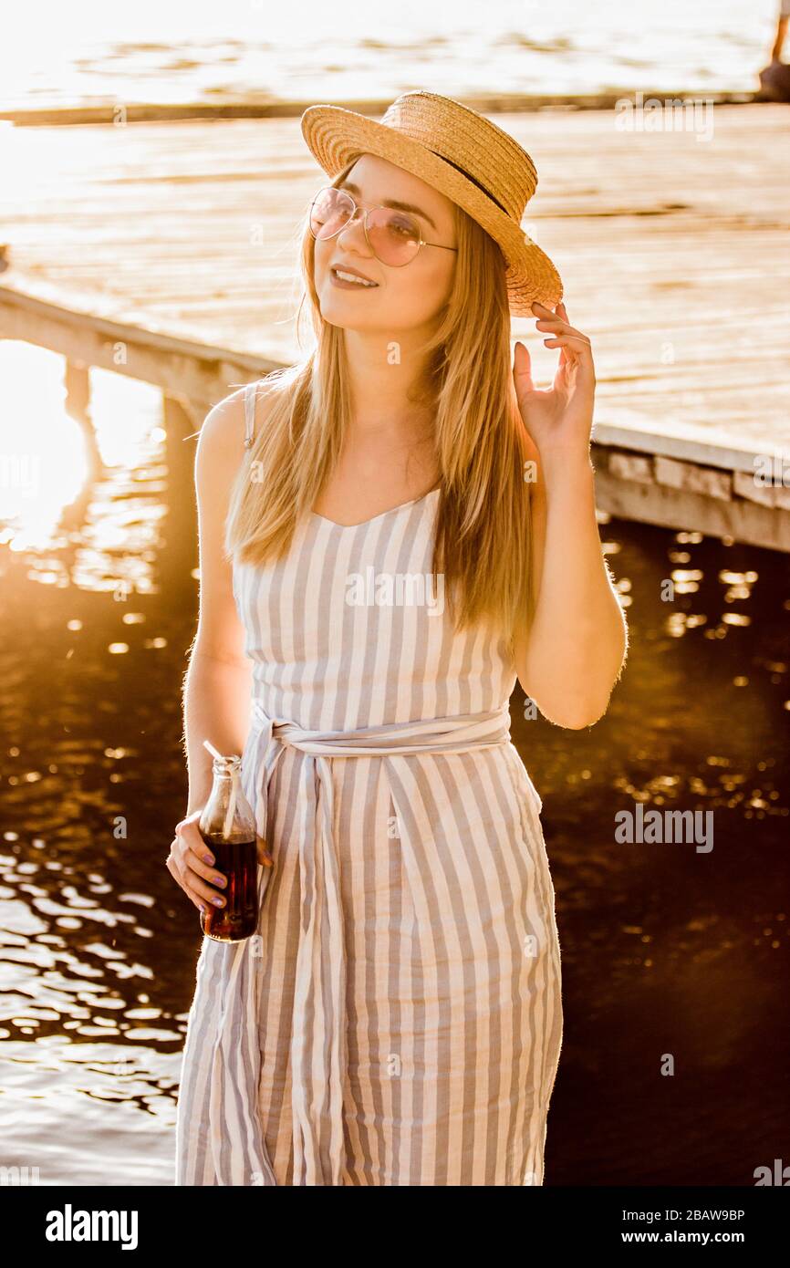 Portrait of happy young woman in straw hat and stripped overalls holding bottle of lemonade in hand while standing at riverside. Beautiful blonde girl Stock Photo