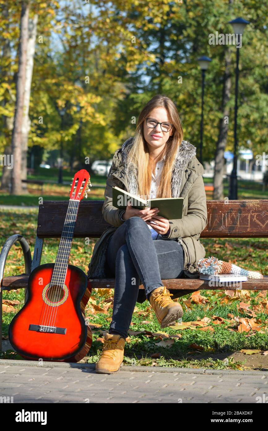 Young, cute girl with spectacles reading a book on the bench in a park, with red acoustic guitar leaning against the bench Stock Photo