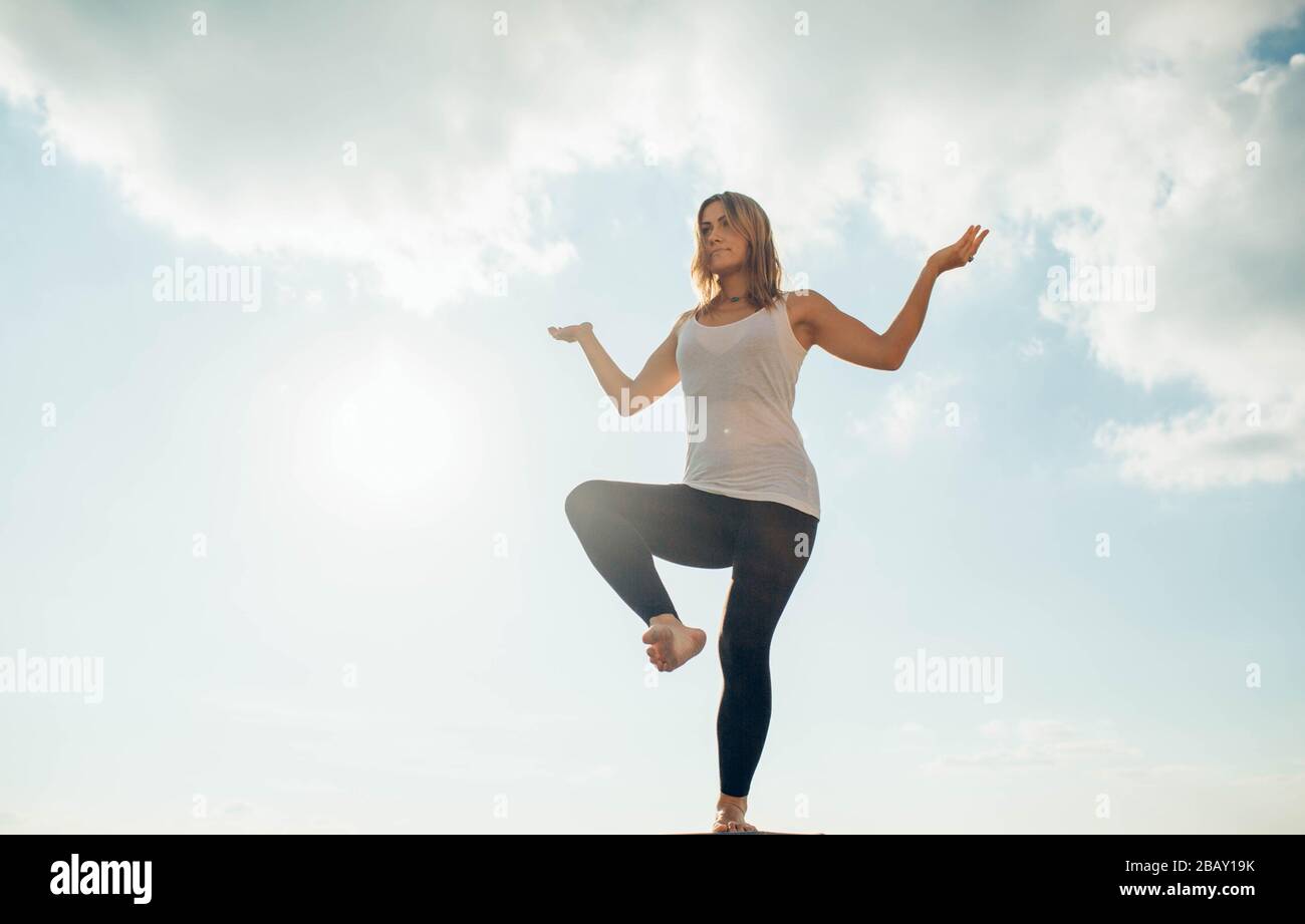 Young woman practices yoga outside. Blonde girl standing on one leg, another raised bent at knee. Hands on sides bent at elbows and turned up. Sky and Stock Photo