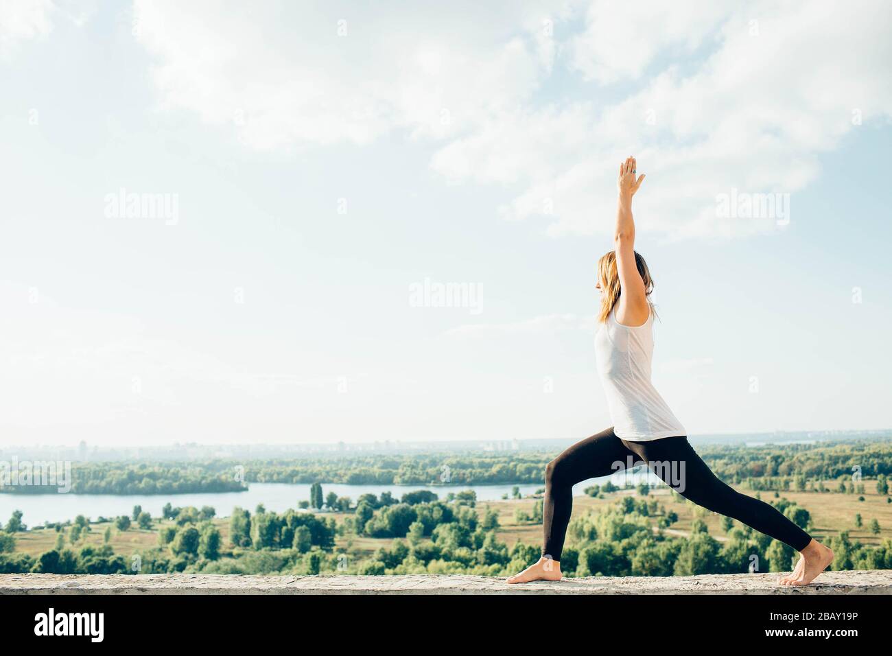 Young woman practices yoga outside. Blonde girl standing in Warrior I posture Virabhadrasana 1 , full length on parapet on beautiful view. Trees river Stock Photo