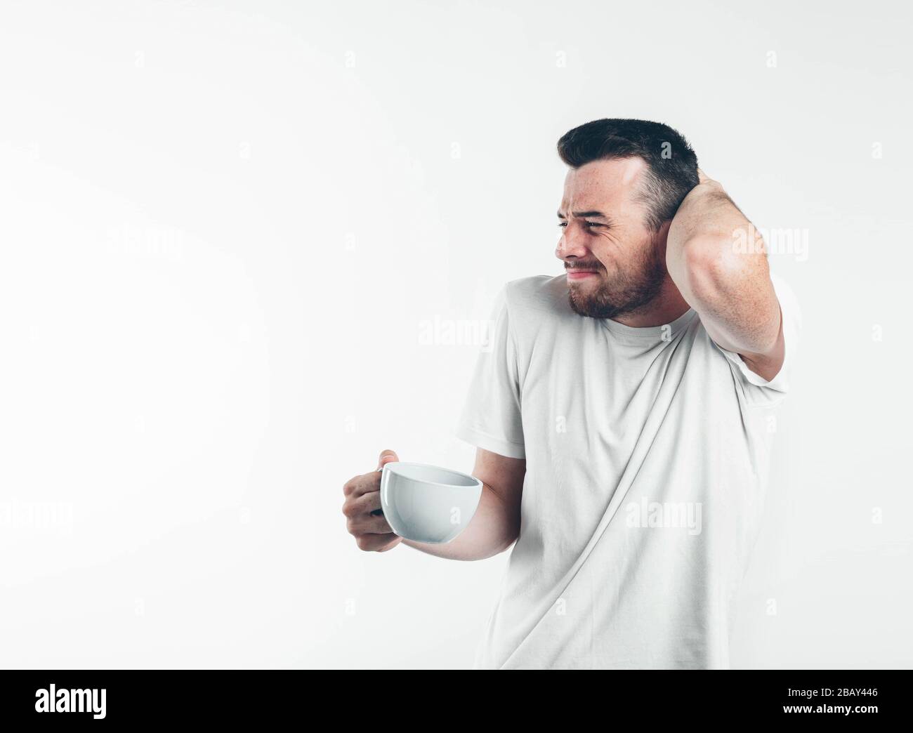 Young brunette man with beard dressed in white T-shirt. Guy touch his occiput by one hand another holds white cup eyes closed. He looks ill. Isolated Stock Photo