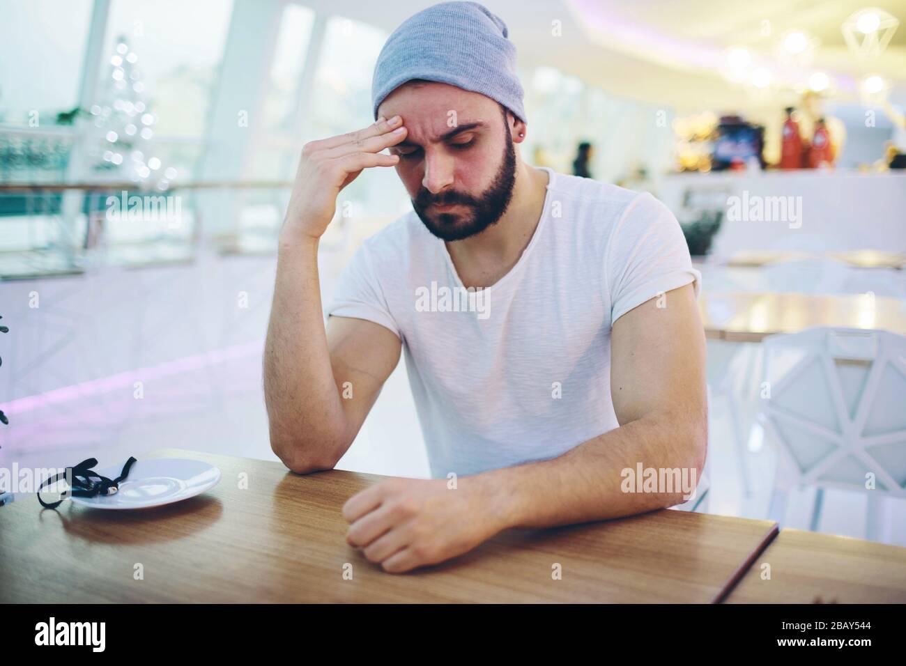 Tired young man has headache. Sit in cafe and hold hand on head. Hipster caught cold or pain in head. Sit inside restaurant or odern cafe. Stock Photo