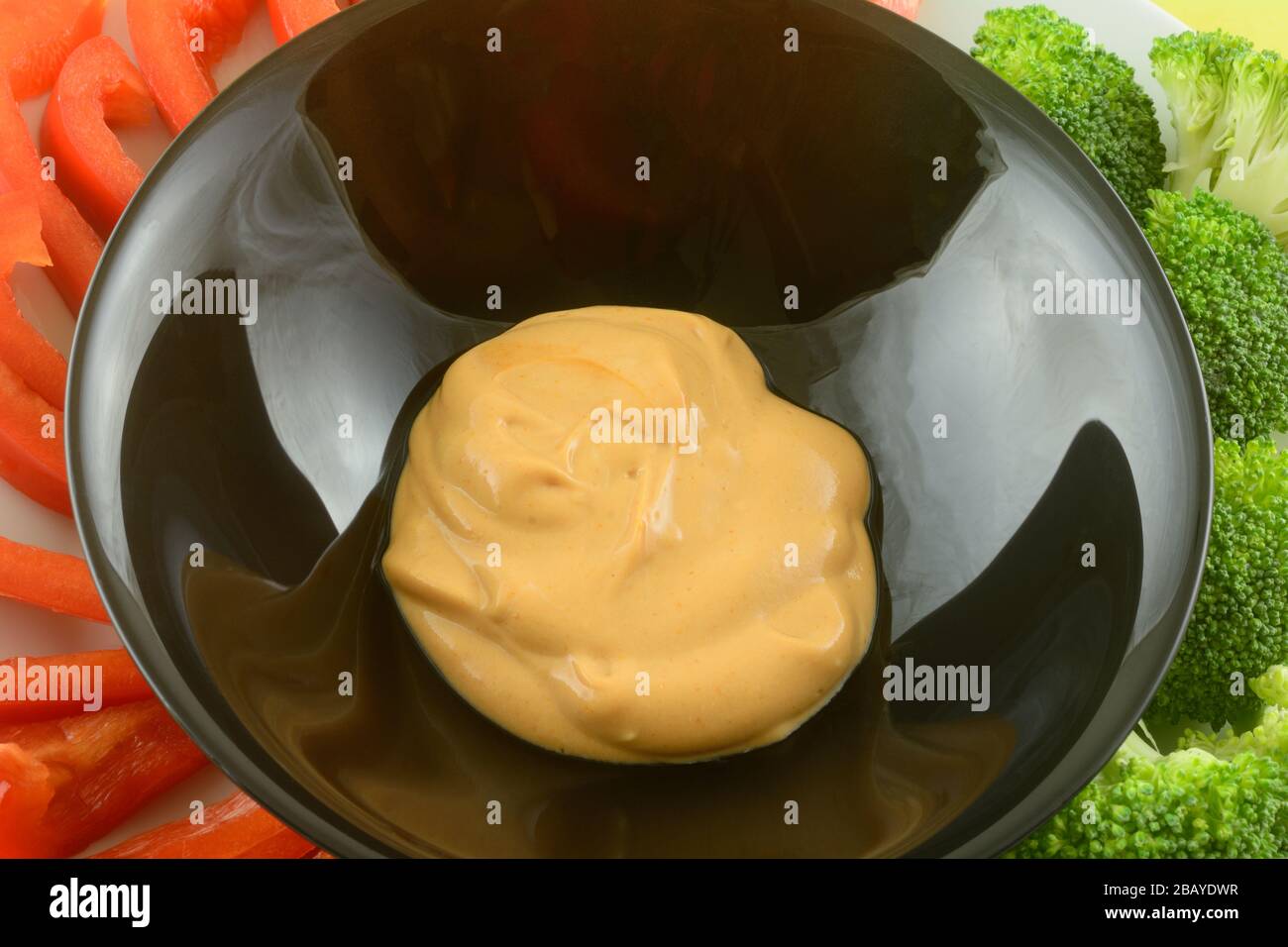 Creamy dip in black bowl with fresh raw red pepper slices and broccoli florets on plate Stock Photo